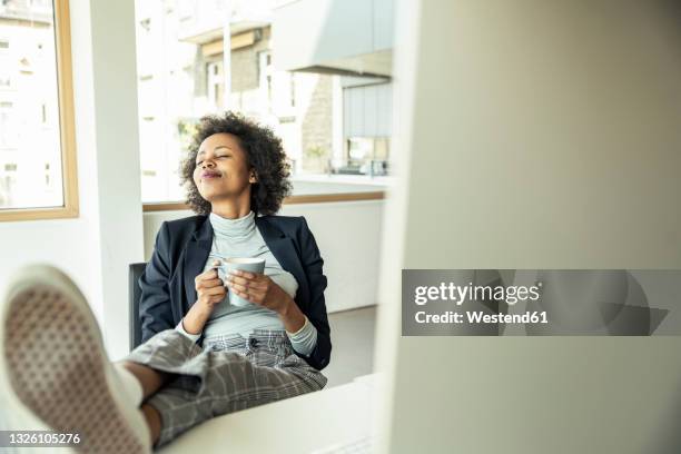 female entrepreneur holding coffee cup while relaxing at office - feet on table stock pictures, royalty-free photos & images