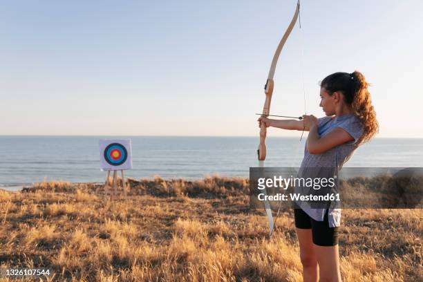 archeress aiming at target while standing on grass during sunny day - archery target stock pictures, royalty-free photos & images
