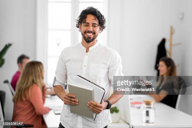 smiling male professional holding book and laptop at coworking office - casual male standing stock-fotos und bilder