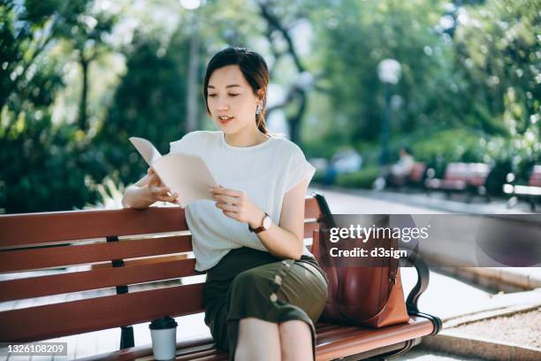 young asian woman sitting on a bench in park, reading a book and having a relaxing time enjoying the sunny day outdoors with coffee in the city. enjoying a technology-free moment - mujer leyendo libro en el parque fotografías e imágenes de stock