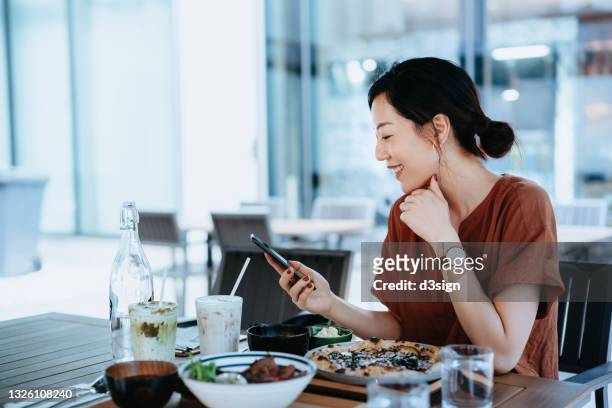 beautiful smiling young asian woman checking social media while having lunch at dining table in outdoor restaurant. enjoying a relaxing afternoon. staying connected on the internet - lunch lady foto e immagini stock