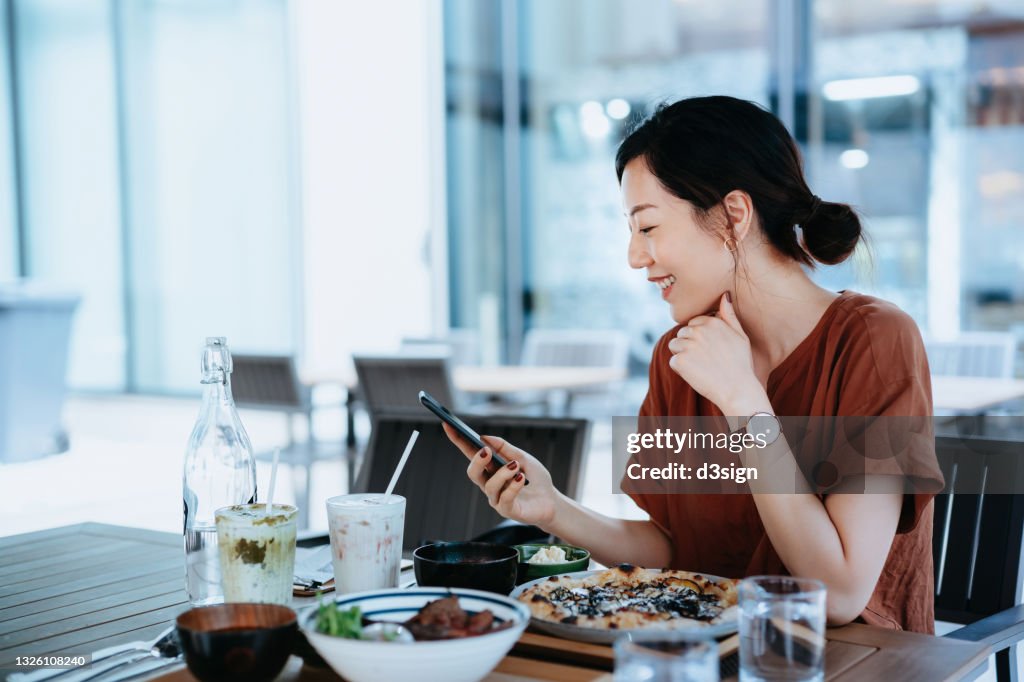 Beautiful smiling young Asian woman checking social media while having lunch at dining table in outdoor restaurant. Enjoying a relaxing afternoon. Staying connected on the internet