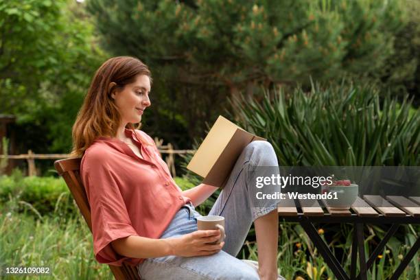 woman with coffee mug reading book while sitting on chair in garden - woman outdoors fotografías e imágenes de stock