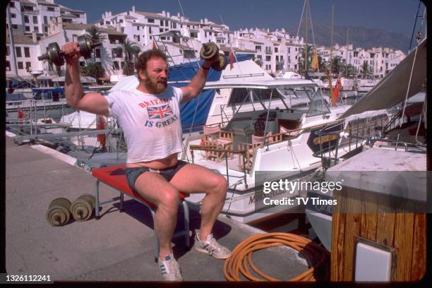 Actor Pat Roach in character as Brian 'Bomber Busbridge lifting weights on the Spanish set of comedy drama Auf Wiedersehen, Pet, circa 1986.