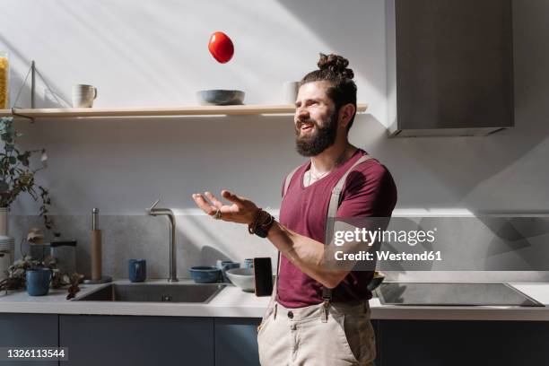 bearded man playing with tomato while standing at kitchen counter - throwing tomatoes stock-fotos und bilder