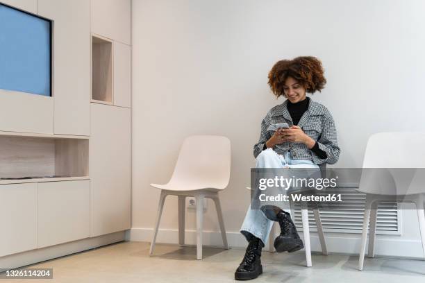 smiling female patient using smart phone while sitting in waiting room at clinic - waiting room clinic stock pictures, royalty-free photos & images