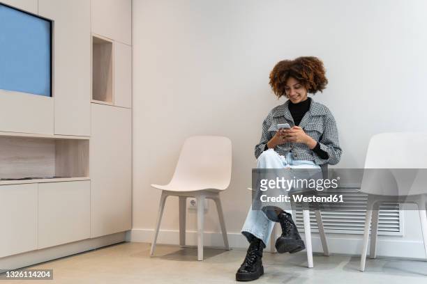 smiling female patient using smart phone while sitting in waiting room at clinic - väntrum bildbanksfoton och bilder