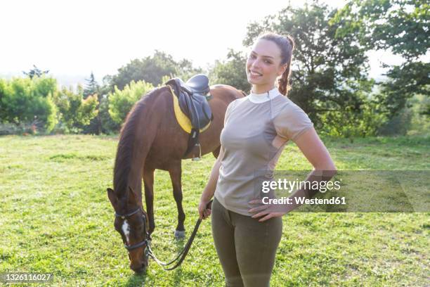 smiling woman holding rein while standing by horse at ranch - grazen stockfoto's en -beelden