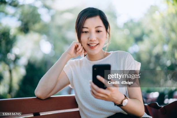 cheerful young asian woman sitting on a bench in park, text messaging on smartphone while having a relaxing time enjoying the sunny day outdoors in the city. lifestyle and technology - donna sorride cellulare foto e immagini stock