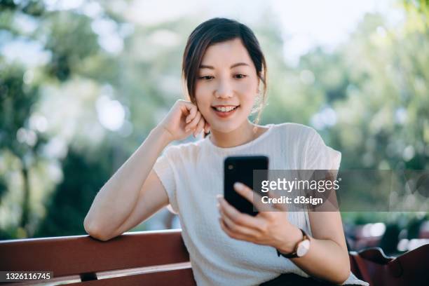 cheerful young asian woman sitting on a bench in park, text messaging on smartphone while having a relaxing time enjoying the sunny day outdoors in the city. lifestyle and technology - asian outdoor ストックフォトと画像