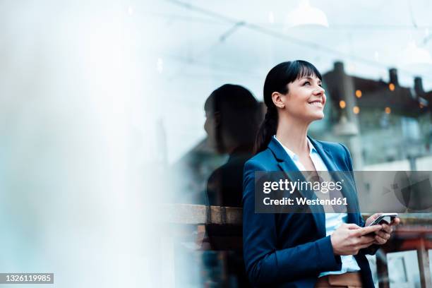 thoughtful businesswoman holding mobile phone while leaning on cafe window - corporate background stock pictures, royalty-free photos & images