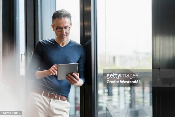 male entrepreneur using digital tablet while leaning on glass window in office - tablet pc stock-fotos und bilder