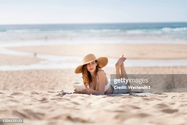 young woman with book looking away while lying on sand at beach during vacations - woman towel beach stock pictures, royalty-free photos & images