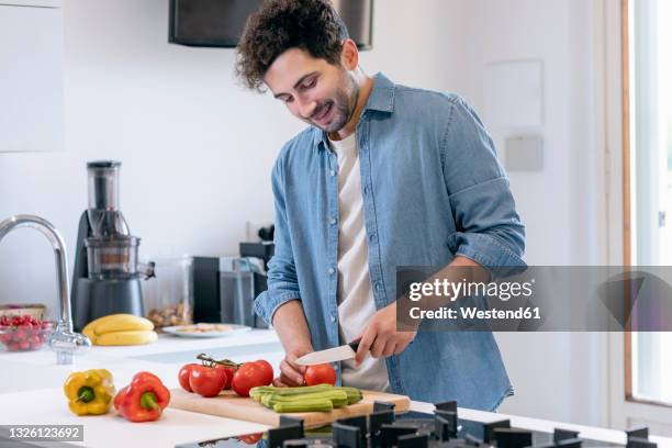 smiling mid adult man cutting tomatoes in kitchen at home - chop stock pictures, royalty-free photos & images