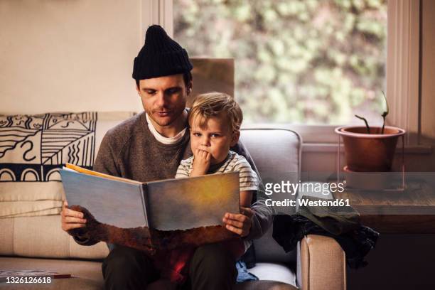 father reading story to son while sitting on sofa at home - storytelling children stock pictures, royalty-free photos & images