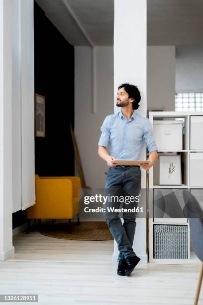 young businessman holding digital tablet while standing in office - legs crossed at ankle stock-fotos und bilder