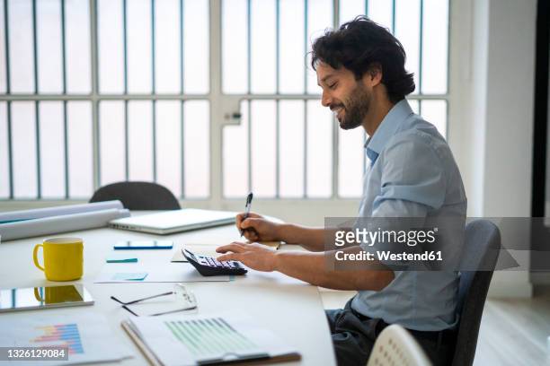 smiling young businessman working while sitting by desk in office - accounting stock pictures, royalty-free photos & images