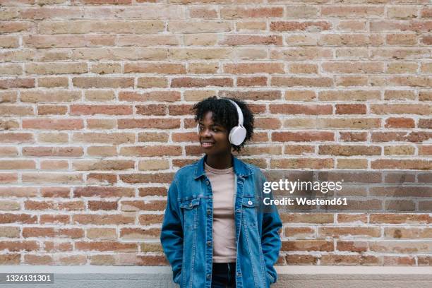 smiling woman with hands in pockets listening music while leaning on brick wall - black hair texture stock pictures, royalty-free photos & images