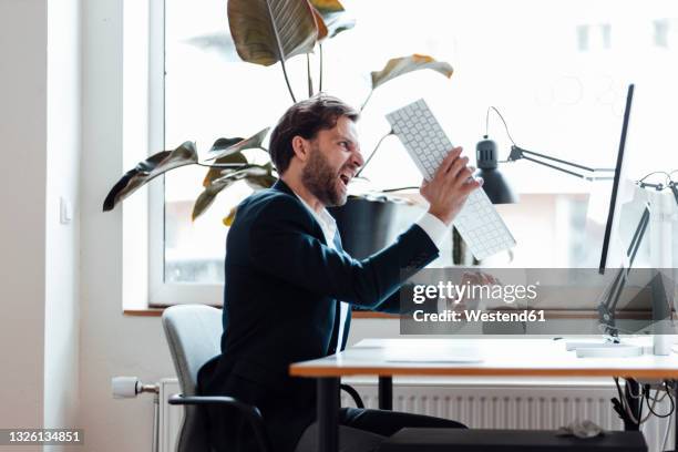 angry businessman holding computer keyboard while sitting by desk - grimm stock-fotos und bilder