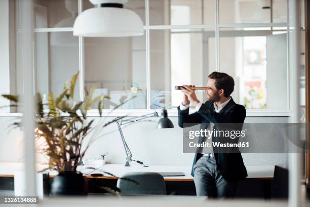 male entrepreneur looking through binoculars while sitting on desk - spy glass businessman stock pictures, royalty-free photos & images