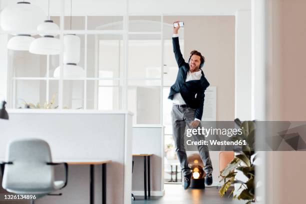 businessman with mobile phone cheering while jumping in office - man mid air stock pictures, royalty-free photos & images