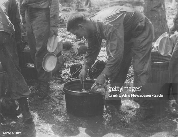 Black and white photograph of a soldier bent over serving food out of a large pot, the pot has been placed on the mud in which the soldier stands,...
