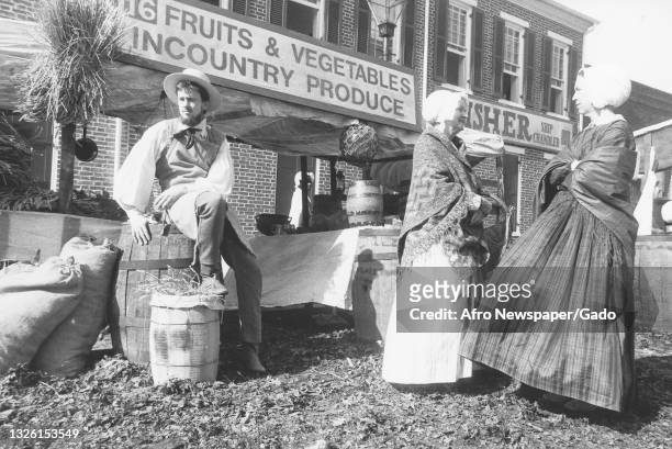 Photograph of three people participating in a reenactment of an 1840's street life scene, a man in period clothing sits atop a barrel in front of a...