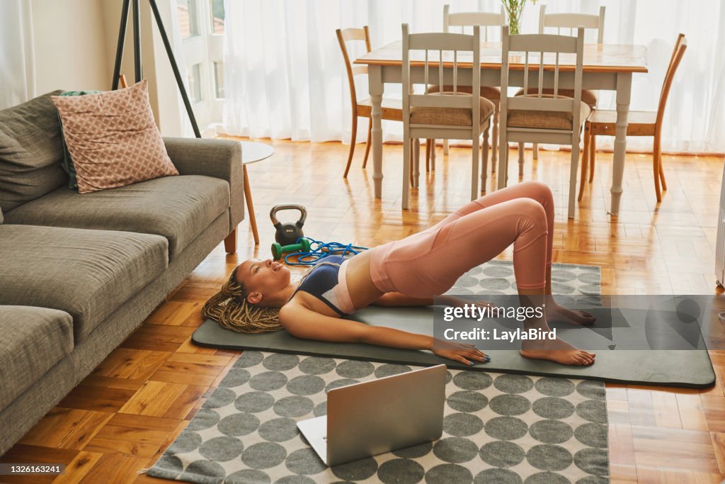 Shot of a young woman using a laptop while doing glute bridge exercises at home