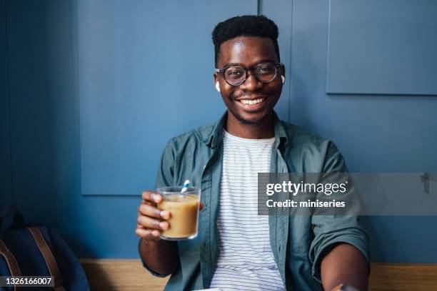 retrato de un hombre de negocios sonriente con auriculares inalámbricos en los oídos sentado en una cafetería y sosteniendo café helado - café con hielo fotografías e imágenes de stock