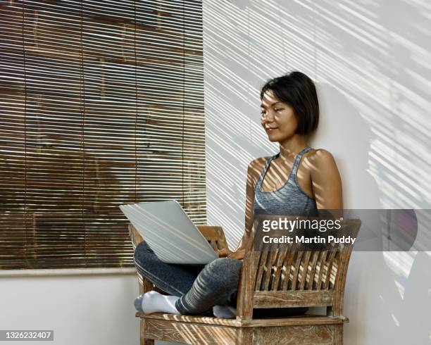 asian woman working from home on open veranda using laptop, with sunlight streaming through bamboo blinds - sportswear stock pictures, royalty-free photos & images