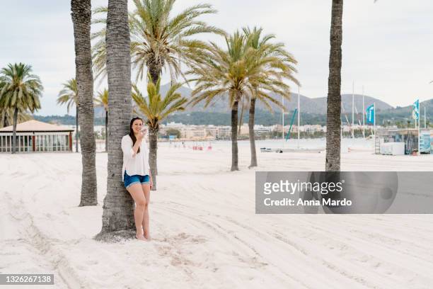 woman talking on her mobile phone at the beach leaning at a palm tree - alcudia stockfoto's en -beelden