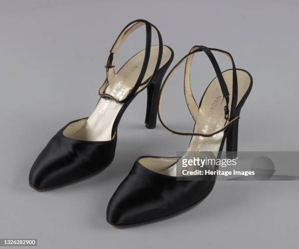 Pair of black satin close-toed shoes with stiletto heels and an ankle strap that is sewn in a cross at the back of the ankle and closes at the...