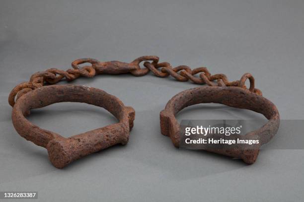Two iron shackles consisting of iron loops joined together by a length of iron chain. The iron is heavily rusted. These would have been used to...