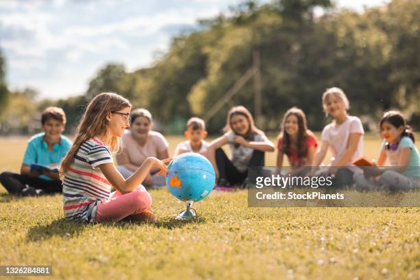 little girl sitting whit her friends on the meadow and pointing to the globe - summer school stock pictures, royalty-free photos & images