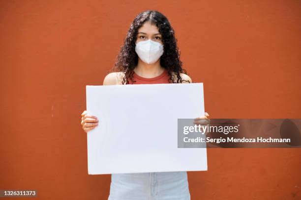 young latino woman with curly hair wearing a face mask and holding a blank sign - person holding blank sign fotografías e imágenes de stock