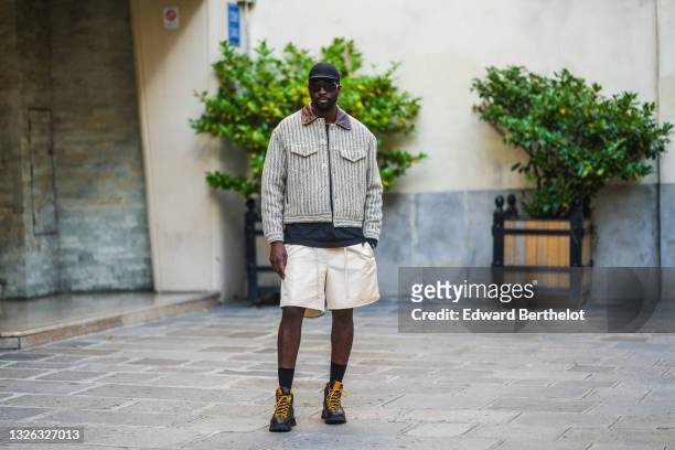 Guest wears a black cap, black sunglasses, a black t-shirt, a dark and pale gray ribbed shirt jacket with brown shiny leather collar, beige shorts,...