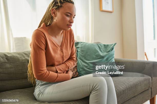 colpo di una giovane donna che prova mal di stomaco mentre giace sul divano di casa - toccare foto e immagini stock