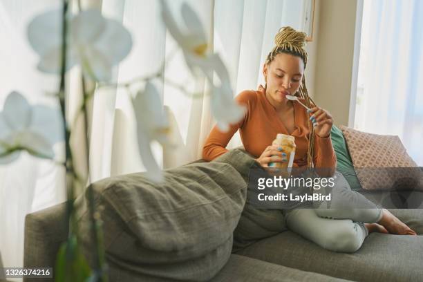 shot of a young woman snacking on peanut butter on the sofa at home - pindakaas stockfoto's en -beelden