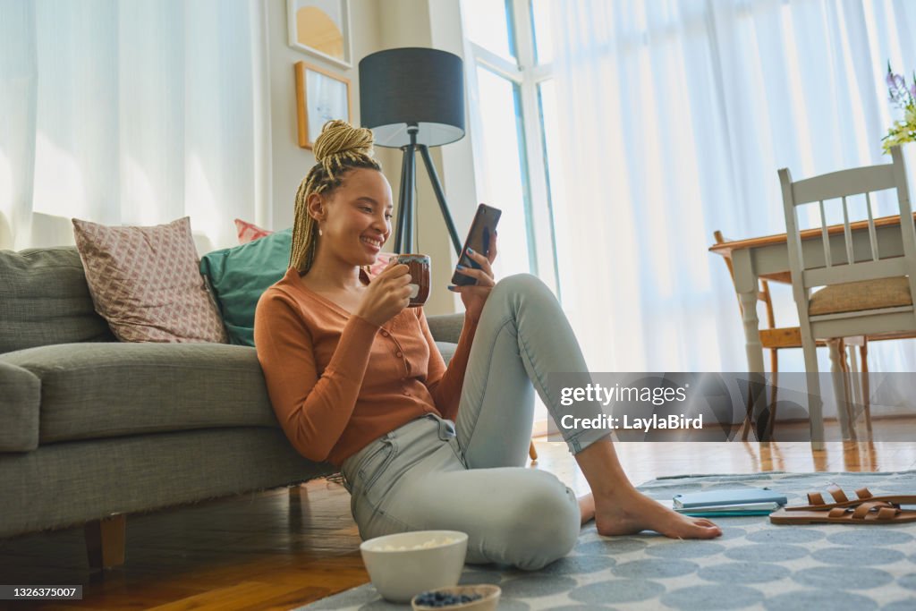 Foto de una mujer joven usando un teléfono inteligente y tomando café mientras trabajaba desde casa