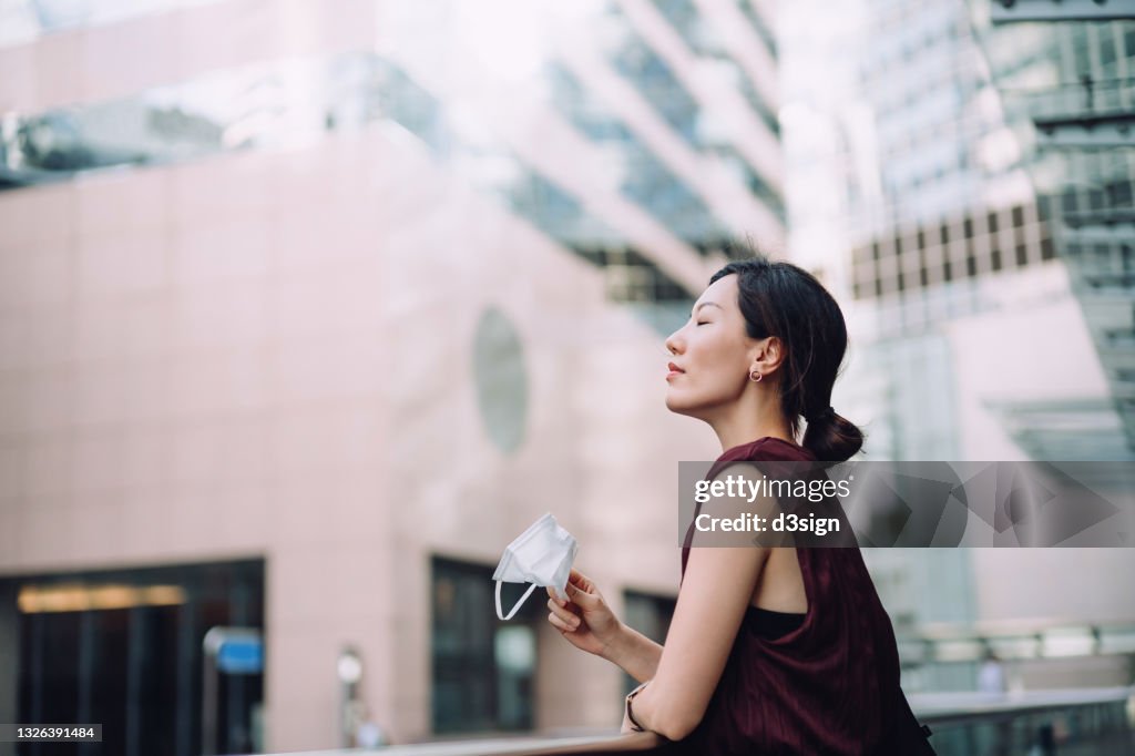Young Asian woman taking off protective face mask, setting herself free and feeling relieved. Head up with her eyes closed. Taking a break from busy and stressful work life in downtown district in the city