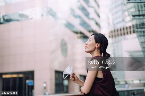 young asian woman taking off protective face mask, setting herself free and feeling relieved. head up with her eyes closed. taking a break from busy and stressful work life in downtown district in the city - end of life care stock pictures, royalty-free photos & images