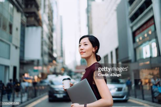 confident and modern young asian businesswoman holding laptop, commuting to work in central business district, against urban skyscrapers and busy traffic in the city. female leadership, determined to success. business on the go - escritório tecnologia olhar em frente imagens e fotografias de stock