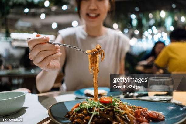close up of joyful young asian woman eating freshly served authentic malaysian-style flat rice noodle in a fusion restaurant. an iconic taste of local malaysian cuisine. asian style cuisine and culture. eating out lifestyle - vietnamese food stockfoto's en -beelden