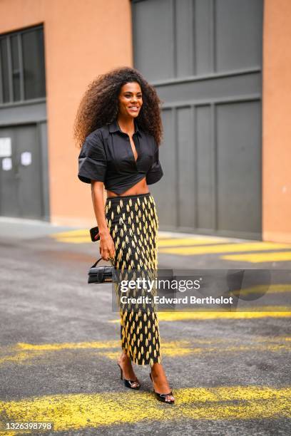 Tina Kunakey wears a black cropped shirt, a black high-waist long skirt with yellow attached sticks, a black mini leather Jacquemus bag, black high...