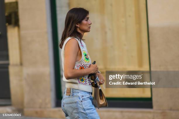 Guest wears a gold earring, a beige wool with pale multicolored print pattern sleeveless t-shirt, blue faded denim mom jeans pants, a brown shiny...