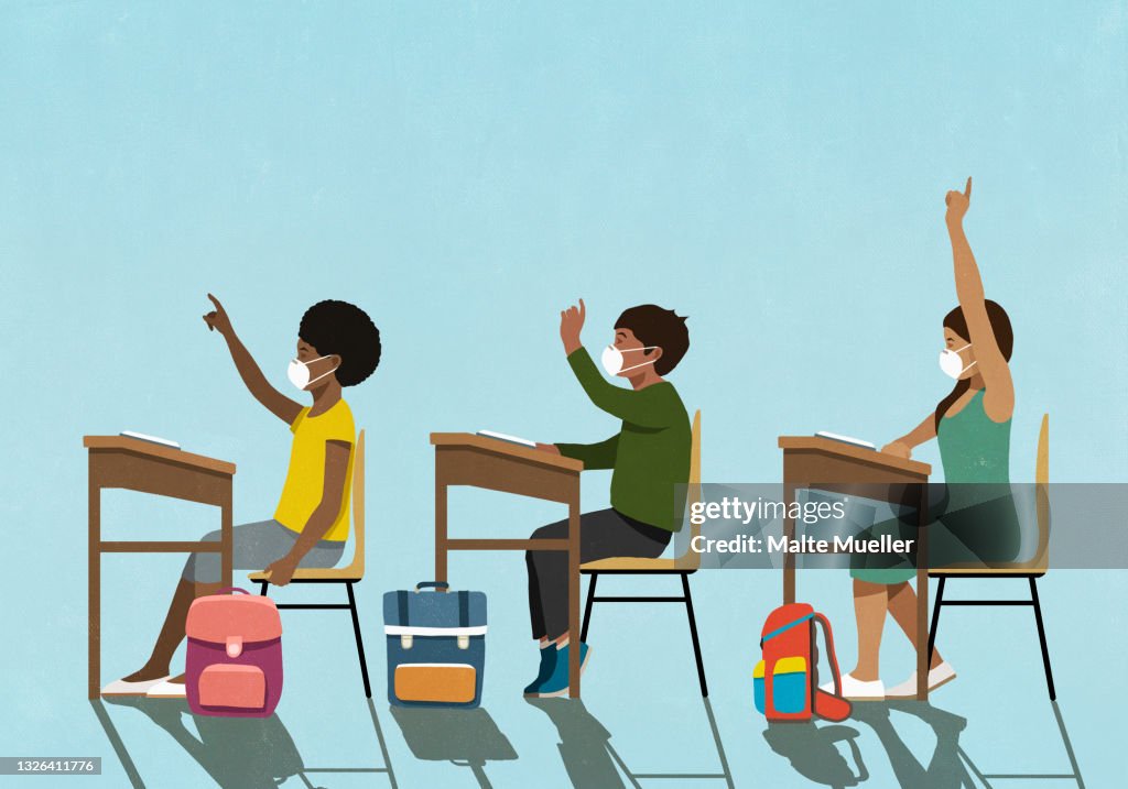 School children in face masks raising hands at classroom desks