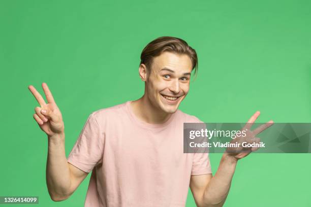 portrait playful young man gesturing peace sign on green background - portrait playful caucasian man foto e immagini stock