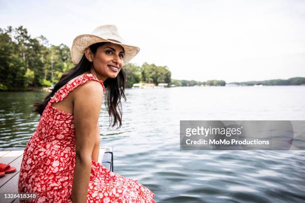 portrait of beautiful woman sitting on dock at lake - chapéu vermelho imagens e fotografias de stock