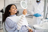 Happy Black Female Patient Looking At Mirror After Dental Treatment In Clinic