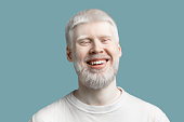 Portrait of happy bearded albino man with pale skin and white hair smiling to camera on turquoise background